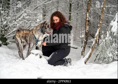 Young woman is kneeling next to her akita inu dog with gray fur, looking at her dog and smiling, in the forest during winter with lots of snow Stock Photo