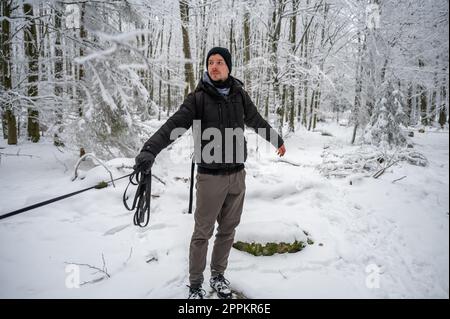 Man with beard and dog leash is standing in the forest during winter with lots of snow, dog running away, master looks at his dog Stock Photo