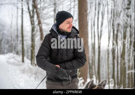 Young man with beard and hat, wearing warm clothes, is smiling, walks his akita inu dog with gray fur in the forest during winter with snow Stock Photo