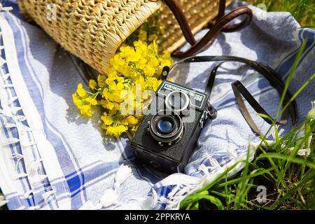 A straw picnic basket lies on a blue blanket on green grass along with a bouquet of yellow flowers. In the background is an old camera with the name Lover 166 written on it. Stock Photo