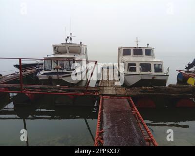 Belgrade, Serbia, January 14, 2020 Two white boats at a wooden pier, left for the winter. Foggy weather in southern Europe. January cloudy day. Reflection of water transport in a river river Stock Photo