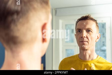 Middle aged man apply moisturizing cream on clear facial areas. Male portrait in room mirror. Skincare morning routine. Stock Photo