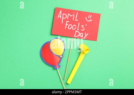 Sign with phrase Happy Fools' Day, paper balloons and party blower on green background, flat lay Stock Photo