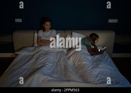 Indifferent man husband using smartphone, ignoring sad unhappy wife while lying together in bed Stock Photo