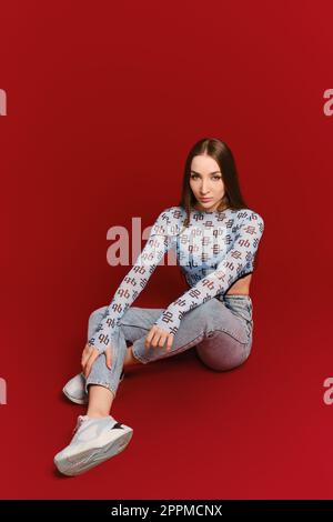Young woman in tank top and jeans posing on the floor of red studio Stock Photo