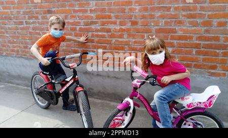 Children 6 and 7 years old in white protective surgical masks go in for cycling. Pause in cycling. A girl in a pink T-shirt and a blond boy in orange. Children on the street near the old brick fence. Stock Photo