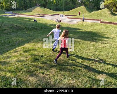 Sremska Mitrovica, Serbia - April 24, 2020- Children run in the park. A boy in a white T-shirt and blue trousers. Girl in a red tank top and trousers. Green lawn or field. People walk in a city park Stock Photo