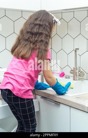 Little girl in protective gloves wash up dishes with sponge in kitchen sink standing on chair. Tidying up kitchen. Stock Photo