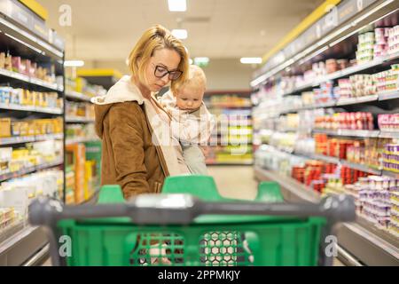 Mother shopping with her infant baby boy child, pushing shopping cart down department aisle in supermarket grocery store. Stock Photo