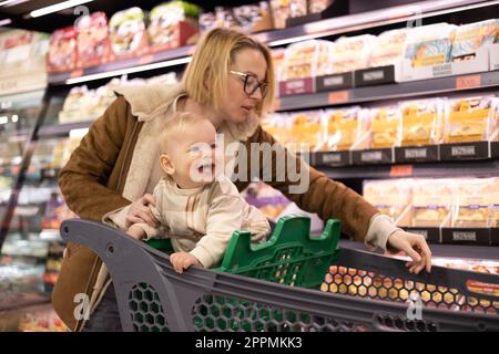 Caucasian mother shopping with her infant baby boy child choosing products in department of supermarket grocery store. Stock Photo