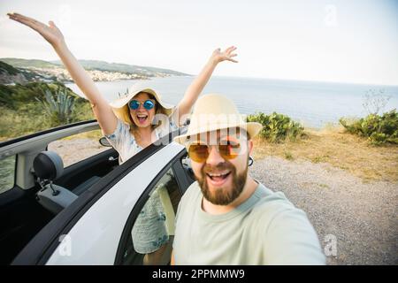 Romantic couple making selfie on smartphone camera in rental cabrio car on ocean or sea beach enjoying summer vacation together and taking picture on cellular resting near sea on weekends Stock Photo