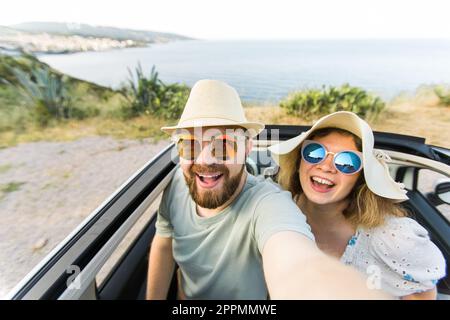 Romantic couple making selfie on smartphone camera in rental cabrio car on ocean or sea beach enjoying summer vacation together and taking picture on cellular resting near sea on weekends Stock Photo