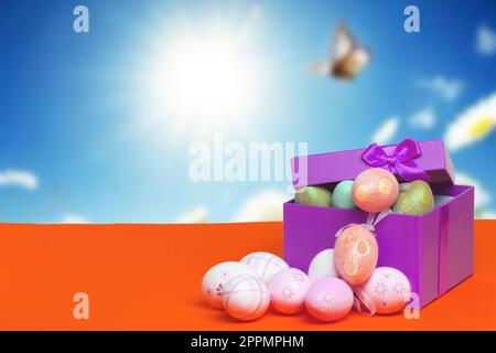 Happy Easter greeting card template. Close up of colourful Easter eggs and an open gift box with eggs on orange table against abstract blurred sunny sky background. Copy space for design. Stock Photo