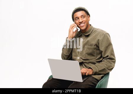 Smiling black freelancer using laptop and talking on mobile phone sitting isolated on white background, african man using computer and having pleasant phone conversation, salesman working remotely Stock Photo