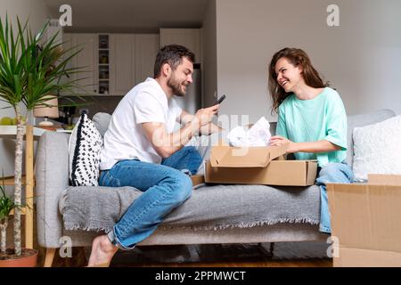 Young married couple sitting on sofa at home and unpacking cardboard boxes with their online purchase. Stock Photo