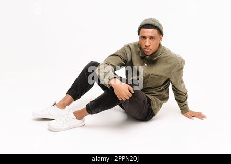Full length photo of fashionable serious handsome young African American man hat and casual shirt sitting on the floor, attractive stylish guy looking at the camera isolated on white Stock Photo