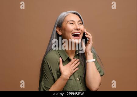 Friendly hilarious mature asian woman talking casually with friend via smartphone, satisfied good mobile connection gray-haired lady holding phone call, enjoying chatting by phone, laughing isolated Stock Photo