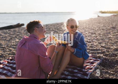 Happy young couple dating together in beach, sitting on a blanket and eating corn. Enjoying holiday time together. Stock Photo