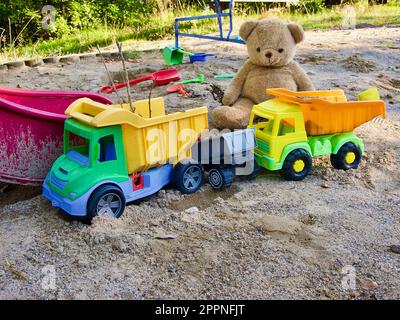 Plastic toy vehicles and a teddy bear in the sandbox at childrens playground outdoors in summer. Stock Photo