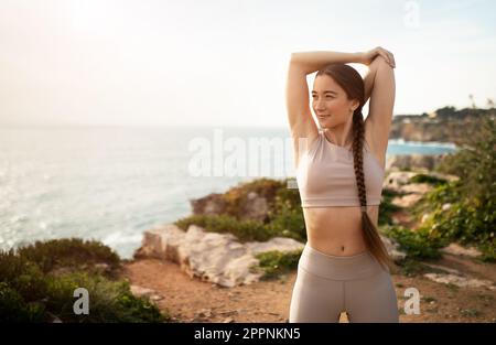 Smiling young caucasian woman in sportswear doing stretching exercise for arms, looks at ocean beach Stock Photo