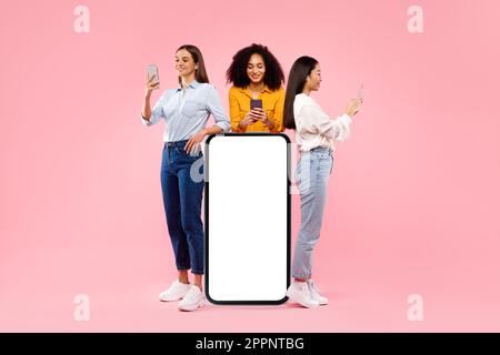 Great app. Multiracial women standing near smartphone with blank white screen using phones, mockup, full body length Stock Photo