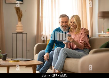 Cheerful Mature Couple Using Digital Tablet Relaxing Sitting At Home Stock Photo
