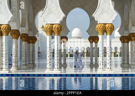Visitors walk through the ornate colonnade of Sheikh Zayed Grand Mosque featuring white marble, gold accents, arches, and domes reflected in water Stock Photo