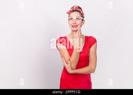 Portrait of attractive charming woman wearing red dress and head band standing looking away, dreaming about future, holding chin. Indoor studio shot isolated on gray background. Stock Photo