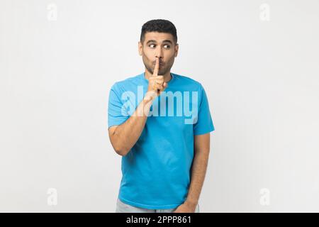 Portrait of funny positive unshaven man wearing blue T- shirt standing looking away, keeps finger near lips, promise to keep secret. Indoor studio shot isolated on gray background. Stock Photo