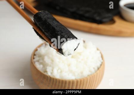 Chopsticks with cooked rice wrapped in nori sheet over white table, closeup Stock Photo