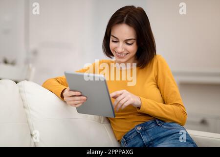 Smiling young woman sitting on couch at home, using pad Stock Photo