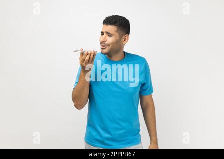 Portrait of handsome unshaven man wearing blue T- shirt standing holding smart phone, recording voice message, or using voice assistant. Indoor studio shot isolated on gray background. Stock Photo