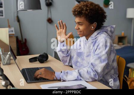 Happy schoolboy in hoodie greeting teacher on laptop screen by waving hand in the beginning of online lesson while sitting in bedroom Stock Photo