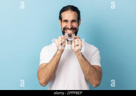 Portrait of man with beard in white T-shirt using teeth whitening braces, dental aligner retainer, dental clinic for beautiful teeth, treatment course. Indoor studio shot isolated on blue background. Stock Photo