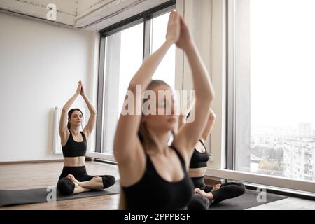 Relaxed caucasian women in cozy activewear performing yoga workout with hands high above heads in anjali mudra in meditation room. Padmasana exercise stretching body and calming mind during session. Stock Photo