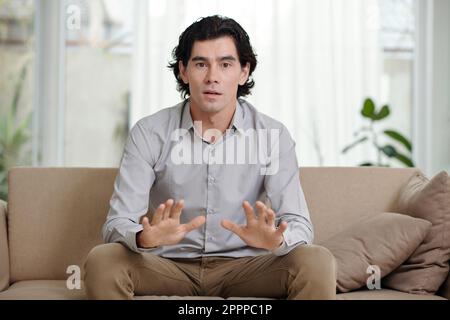 Young confident counselor sitting on couch in front of camera and explaining something while consulting online patient Stock Photo