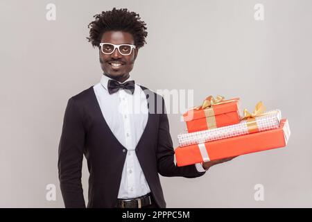 Portrait of joyful cheerful man in glasses standing holding three present boxes, being in festive mood, wearing white shirt and tuxedo. Indoor studio shot isolated on gray background. Stock Photo
