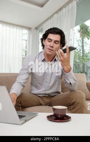 Confident young man with smartphone talking on speakerphone and typing on laptop keyboard while sitting on couch by his workplace Stock Photo