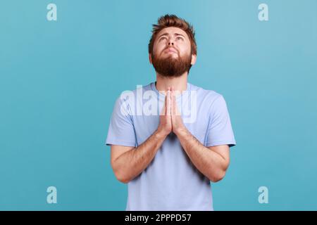 Portrait of hopeless desperate bearded man folding hands in pray looking up, communicating with god, asking for forgiveness and blessing. Indoor studio shot isolated on blue background. Stock Photo