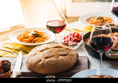 Table set for dinner with the sunset light filtering through. Plates with spaghetti with tomato sauce and Italian pasta, bread and glasses with red wi Stock Photo