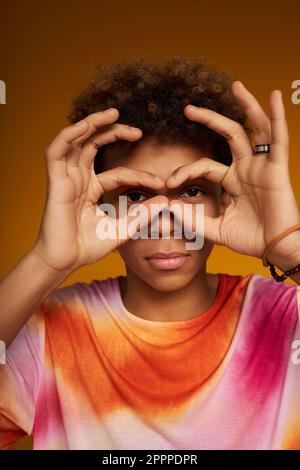 Close-up of teenage boy looking at camera keeping hands by his eyes during photo shooting against yellow background in isolation Stock Photo
