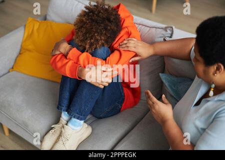 Mature woman sitting on couch next to depressed teenage guy in casualwear and touching his shoulder while comforting him Stock Photo