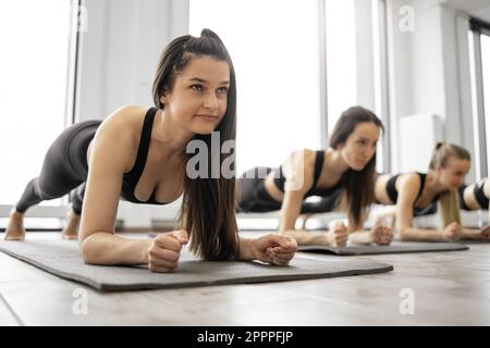 Athletic young females strengthening whole body with Forearm Plank Pose during yoga workout practice in fitness center. Sporty women in activewear improving posture with Phalakasana II exercise. Stock Photo