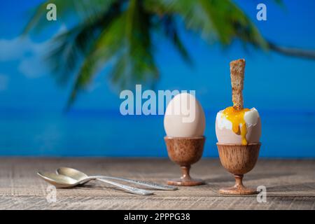 Soft boiled egg in eggcup with slice of toasted toast on wooden table with sea water, coconut palm tree and blue sky background on sunny summer day in Stock Photo