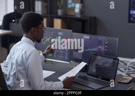 African American broker analyzing financial market while sitting at table in front of monitors with charts in office Stock Photo