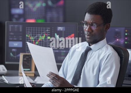 African American businessman in eyeglasses examining financial document while sitting at table with computer monitors with charts in background Stock Photo