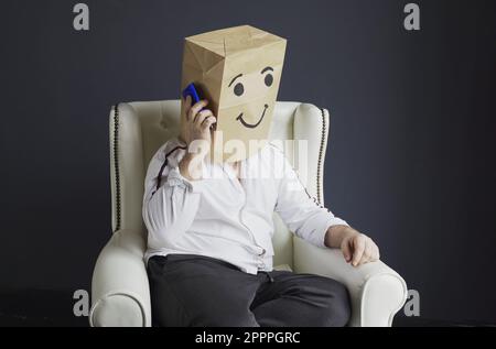 A man in a white shirt with a paper bag on his head, with a smiley face drawn, talking on the phone. Stock Photo