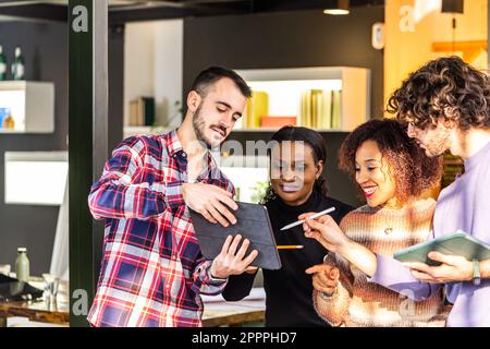 Team of young engineers discussing digital blueprint on note pad at meeting. Smiling people and sunset light on a coworking space. Stock Photo
