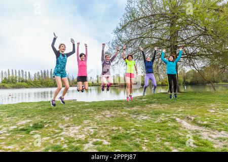 Mixed age group of female runners having fun during a workout. Jump with arms raised in a park near a pond in nature. Wellness active life concept Stock Photo