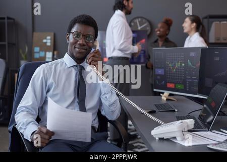African American trader looking at camera while discussing situation on stock market with customer sitting at his workplace Stock Photo